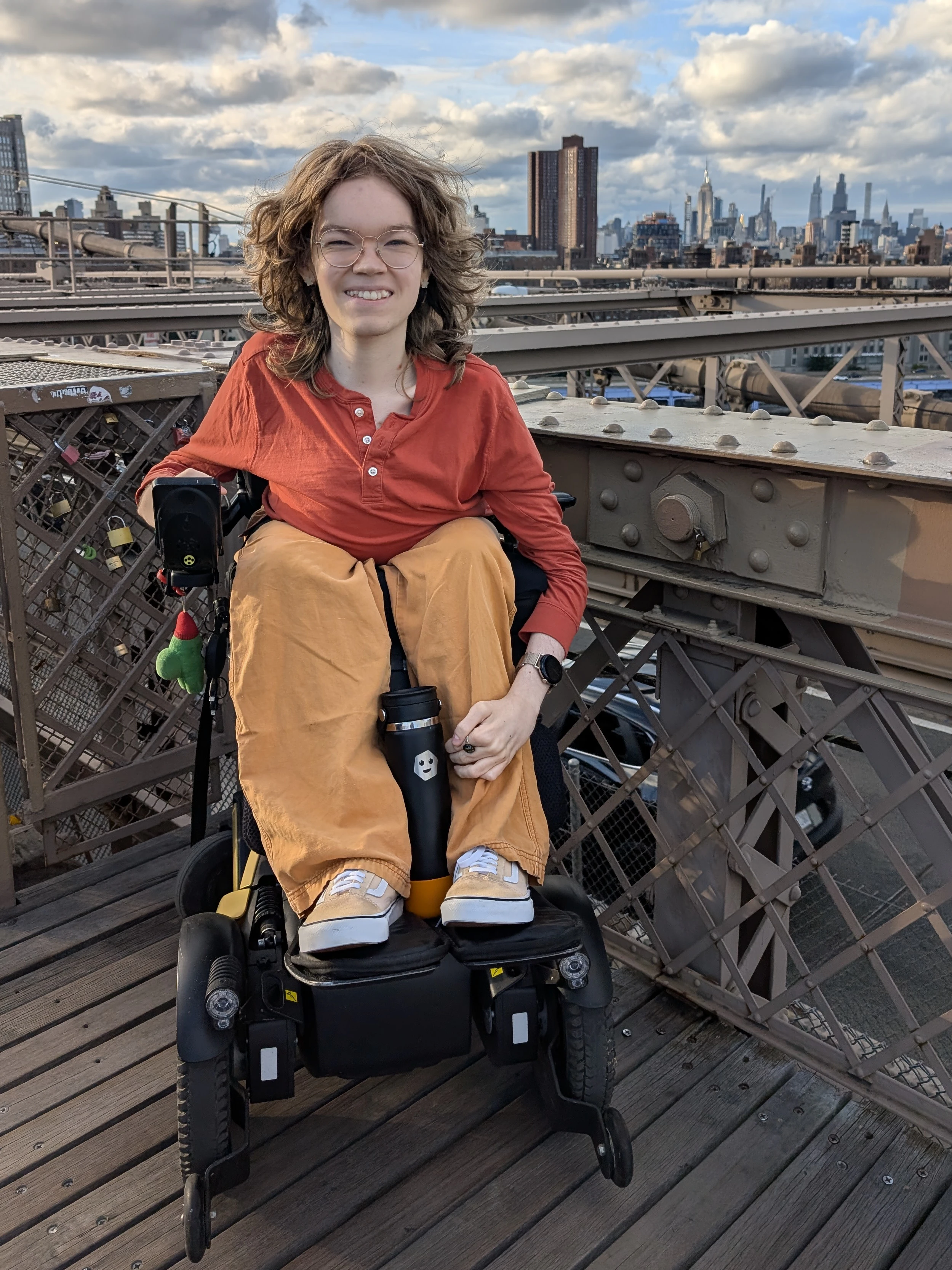 A white androgynous person in a motorized wheelchair. They have medium-long, dirty blonde hair, round gold glasses, and star-shaped earrings. They are smiling.