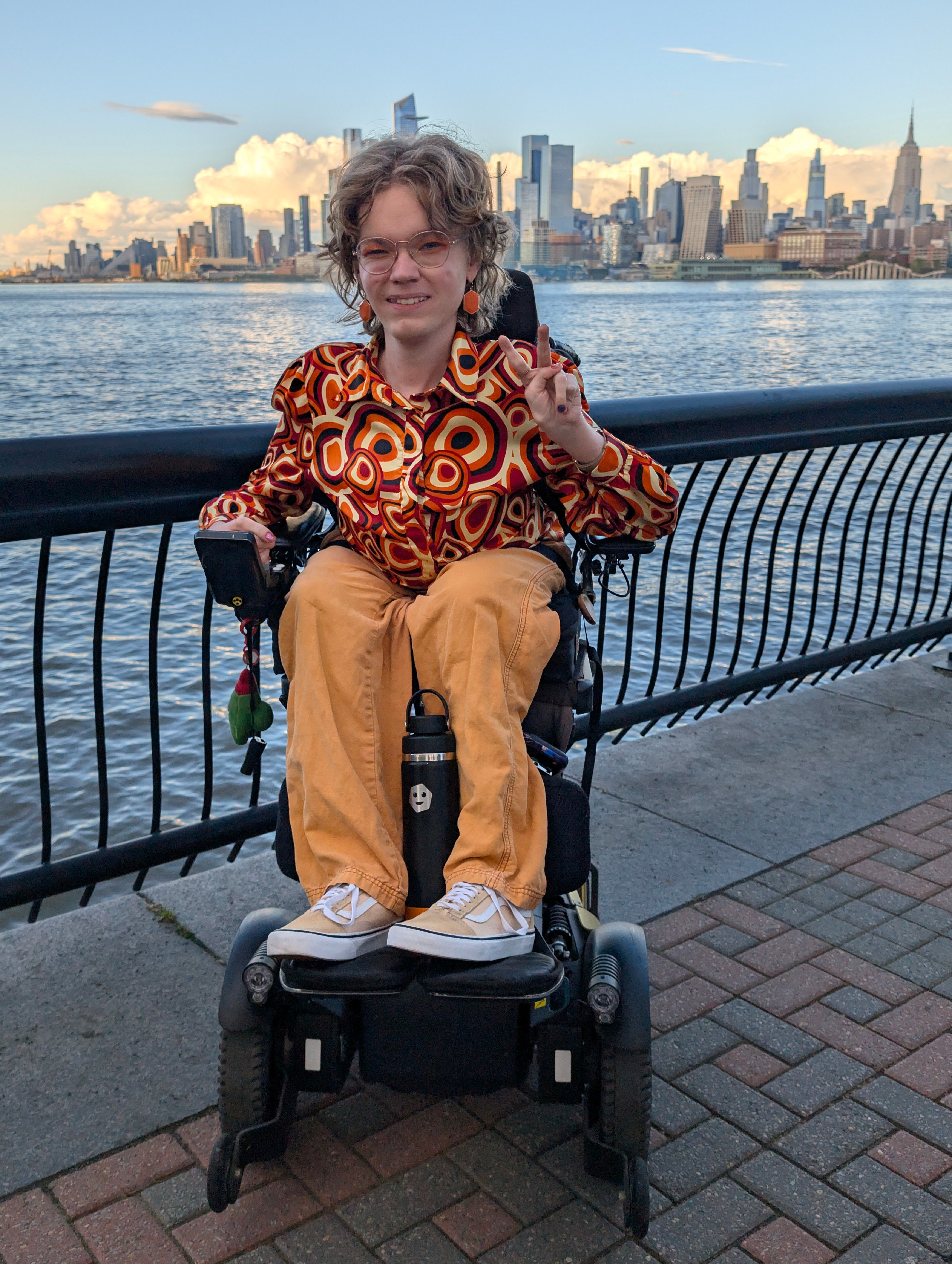 A smiling androgynous person in a motorized wheelchair with their hand making a peace sign. They are wearing all orange. They have medium-long, dirty blonde hair, round gold glasses, and hexagon earrings.